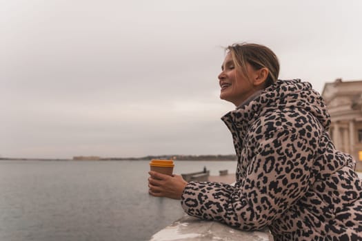 A woman in a leopard print coat is holding a coffee cup and looking out at the water