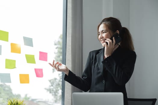 Young creative woman professional using sticky notes in glass wall to writing strategy business plan to development grow to success. Happy asian woman working at office..