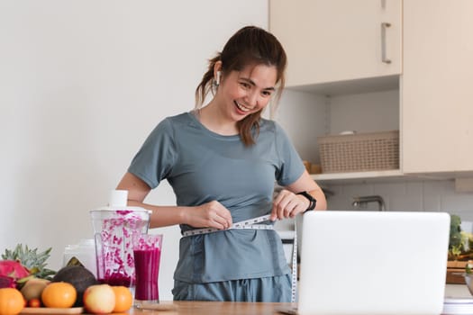 A woman is standing in a kitchen, smiling and holding a measuring tape. She is in front of a blender and a laptop, and there are several fruits and vegetables on the counter