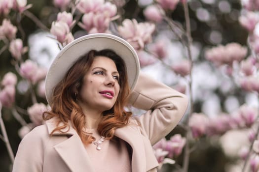 Woman magnolia flowers, surrounded by blossoming trees, hair down, white hat, wearing a light coat. Captured during spring, showcasing natural beauty and seasonal change
