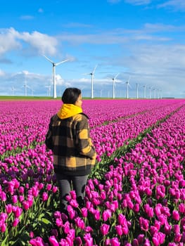 A woman gracefully stands amidst vibrant purple tulips in a picturesque field as the windmill turbines of the Netherlands spin in the background.