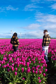 Two individuals standing amongst a field of vibrant purple tulips with windmill turbines in the background during a beautiful Spring day.