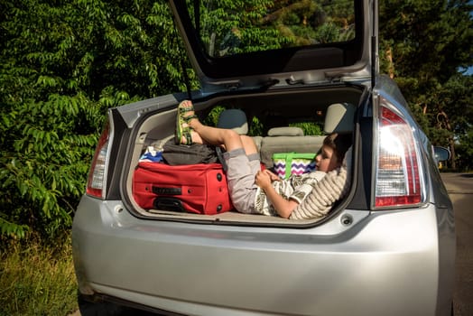 Cute little boy laying on the back of the bags and baggage in the car trunk ready to go on vacation with happy expression. Kid resting playing on smartphone.