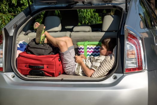 Cute little boy laying on the back of the bags and baggage in the car trunk ready to go on vacation with happy expression. Kid resting playing on smartphone.