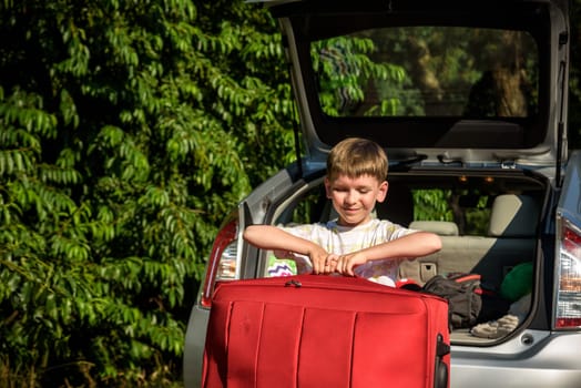 Pretty boy loading the luggage in the trunk of the car. Kid looking forward for a road trip or travel. Family travel by car.