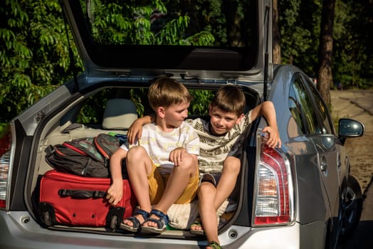 Two cute boys sitting in a car trunk before going on vacations with their parents. Two kids looking forward for a road trip or travel. Summer break at school. Family travel by car.