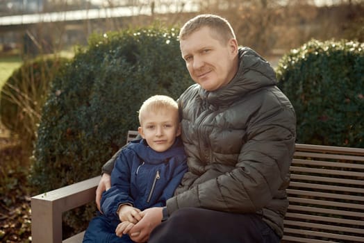 Autumnal Family Affection: Father, 40 Years Old, and Son - Beautiful 8-Year-Old Boy, Seated in the Park. Embrace the warmth of familial love in the autumnal air with this heartwarming image. A father, 40 years old, and his son - a beautiful 8-year-old boy, seated in the park. The essence of autumn adds a touch of seasonal charm to this delightful moment of family affection.