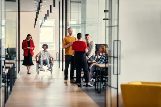 A diverse group of young professionals, including businesswomen and an African-American entrepreneur in a wheelchair, engage in collaborative discussion on various business projects in a modern startup office setting