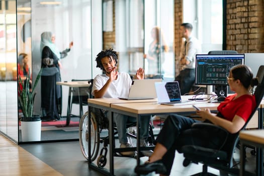 A young business group, including an African American businessman in a wheelchair, collaborates within a modern glass office, actively engaged around a computer and laptop, collectively solving diverse business challenges with determination.