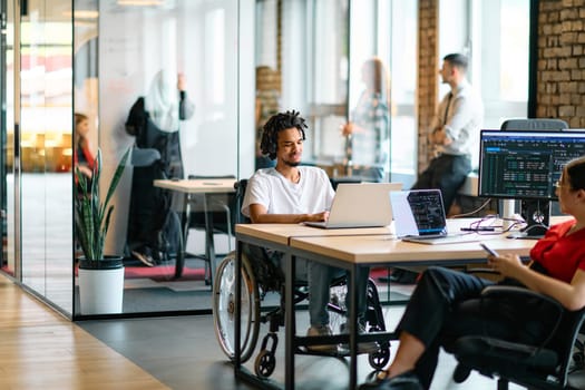 A young business group, including an African American businessman in a wheelchair, collaborates within a modern glass office, actively engaged around a computer and laptop, collectively solving diverse business challenges with determination.