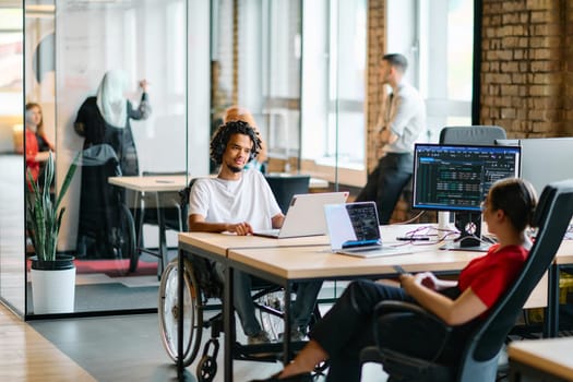 A young business group, including an African American businessman in a wheelchair, collaborates within a modern glass office, actively engaged around a computer and laptop, collectively solving diverse business challenges with determination.