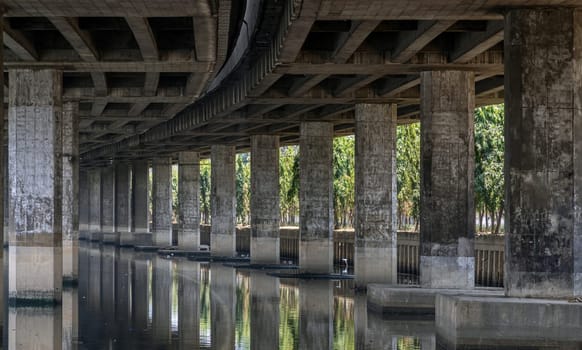 Bangkok, Thailand - Apr 20, 2024 - Perspective view of concrete pillars under Khlong Toei Expressway, with calm surface water reflection wonderful shadow, shape, lines and curve the underside support beams of the road along the Khlong phra khanong canal in Bangkok Thailand, Space for text, Selective focus.