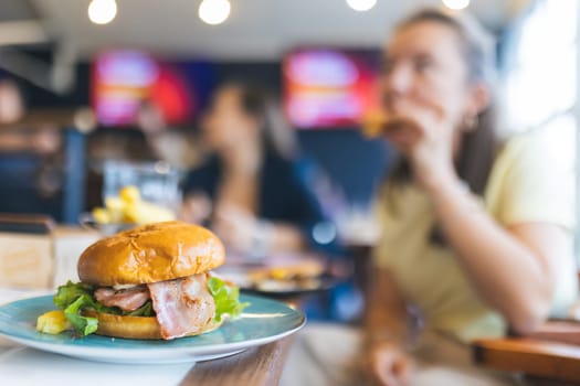 A woman is eating a burger in a restaurant. The burger is on a blue plate and is surrounded by other food items. The woman is looking at the camera, and there are other people in the background