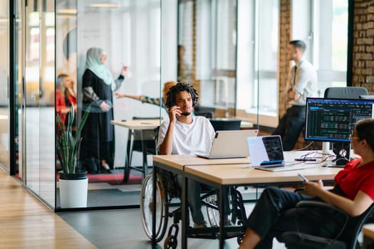 A young business group, including an African American businessman in a wheelchair, collaborates within a modern glass office, actively engaged around a computer and laptop, collectively solving diverse business challenges with determination.