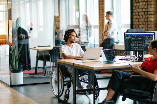 A young business group, including an African American businessman in a wheelchair, collaborates within a modern glass office, actively engaged around a computer and laptop, collectively solving diverse business challenges with determination.