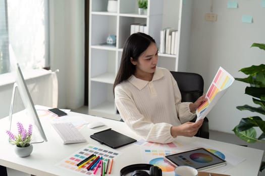 Asian woman freelance graphic designer working with color swatch samples and computer at desk in home office, young lady choosing color gamma for new design project.