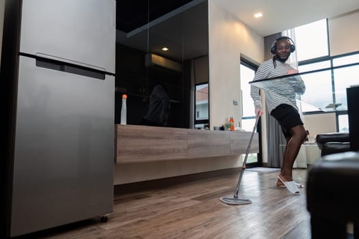 Young African American having fun with mop and music at home, showing dance moves and singing in living room. African American man mopping wooden floors and doing spring cleaning, housework.