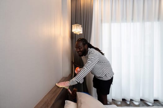 Happy young African American man wear rubber gloves to clean and wipe the headboard of the bed at home.