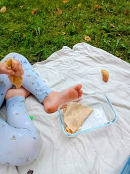 little girl legs in blue pants on linen cloth in fresh spring grass eating pancakes on a picnic. High quality photo Picnic in a forest, on a lawn. Daughter mother picnic together. Eating pancakes