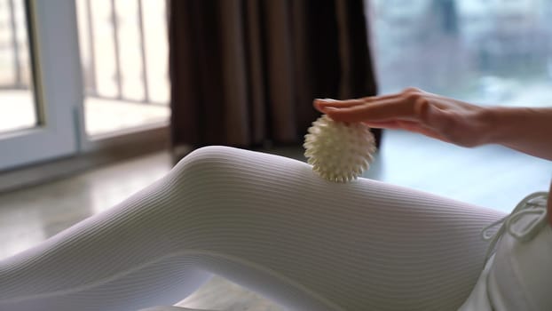 Athletic slim caucasian woman doing thigh self-massage with a massage ball indoors. Self-isolating massage.