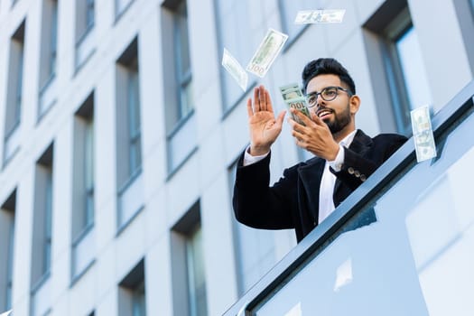 Happy rich Indian businessman wasting throwing money dollars cash around earnings, big profit, win lottery, share, celebrate, charity donation outdoors. Arabian Hindu freelancer guy in downtown city