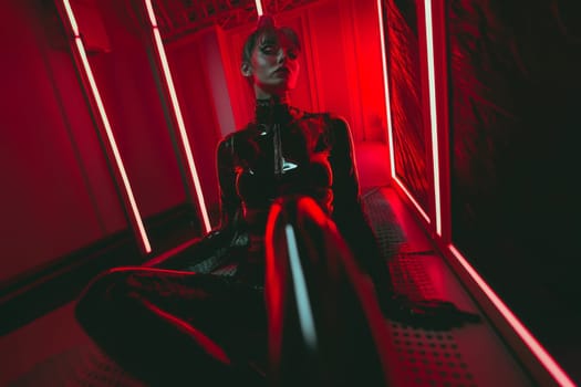 Beautiful young sexy female model in latex sits in a red light in a hangar. Fetish concept