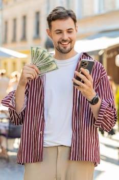 Happy rich bearded man counting holding money dollar cash, use smartphone calculator app, plans to order gifts and food delivery online booking hotel room. Young guy tourist on city street. Vertical