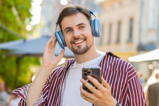 Happy relaxed overjoyed bearded adult man dancing in wireless headphones choosing, listening favorite energetic disco rock n roll music outdoors. Young guy standing on urban city sunshine street
