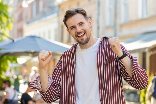Happy joyful bearded adult man shouting, celebrating success, winning, goal achievement, good news, lottery jackpot luck, victory outdoors. Young guy walking in urban city sunshine street. Lifestyles