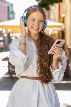 Happy relaxed overjoyed teenager young adult girl in wireless headphones choosing, listening favorite energetic disco rock n roll music dancing outdoor. Child on urban city sunshine street. Vertical