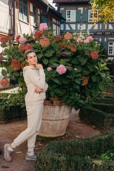 Attractive curly blonde woman walk on the city park street. Girl wear purple hoodie look happy and smiles. Woman make here me gesture standing near pink blooming bush flowers. Happy laughing girl