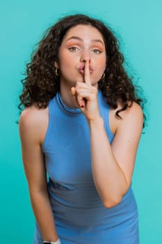 Shh be quiet please. Young woman presses index finger to lips makes silence gesture sign do not tells secret, stop gossip. Pretty brunette adult girl isolated alone on blue studio background. Vertical