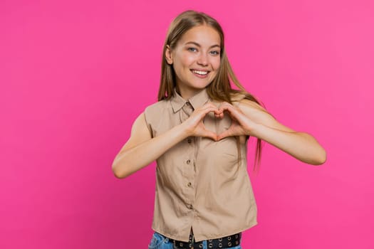 Woman in love. Smiling attractive woman makes heart gesture demonstrates love sign expresses good positive feelings and sympathy. Pretty blonde young girl isolated on pink studio wall background