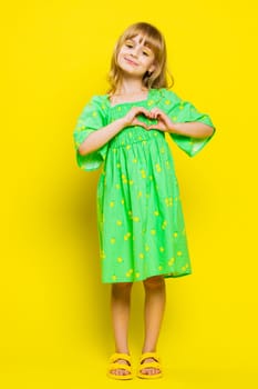 I love you. Smiling blonde school girl makes heart gesture demonstrates love sign expresses good feelings and sympathy. Happy preteen female child kid isolated on studio yellow background. Vertical