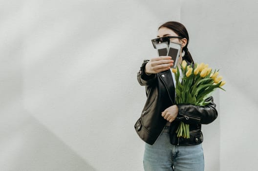 A young beautiful Caucasian brunette girl in sunglasses and a leather kurta with a bouquet of yellow tulips holds out her hand showing two packages of eyelashes for extensions standing on the right near a white wall with copy space on the left on a spring day, side view close-up.