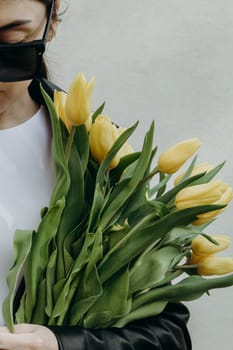 Body part portrait of one young beautiful Caucasian girl in sunglasses leather kurta with a bouquet of yellow tulips standing near a white wall on a spring day and looking down, side view close-up.