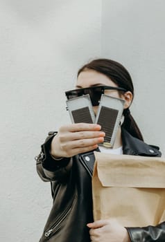 Young beautiful Caucasian brunette woman in sunglasses and leather jacket with paper shopping bag extends her hand showing two packages of eyelashes for extensions standing near a white wall on a spring day, close-up side view with depth of field.