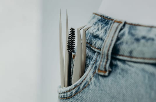 Two different tweezers and a round brush for eyelash extensions protrude from the right pocket of blue jeans, close-up side view with small copy space on the left and depth of field.