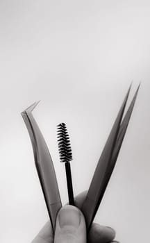 The hand of a young Caucasian girl holds two different tweezers and one round brush for eyelash extensions against a gray-white background, close-up side view. Black and white.