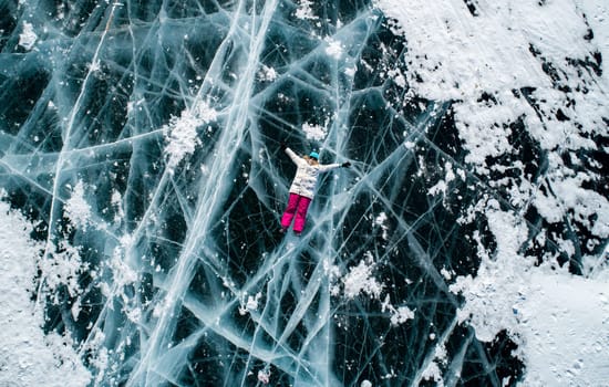 Aerial top down shot of the young woman lying on the blue cracked ice of Baikal and shaking hands to the camera. Popular tourist spot. Winter landscape of frozen Baikal.