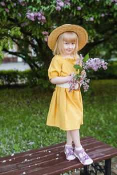 A little girl in a yellow dress and straw hat wearing a bouquet of lilacs. A walk in a spring park, blossoming lilacs