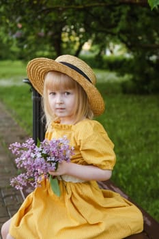 A little girl in a yellow dress and straw hat wearing a bouquet of lilacs. A walk in a spring park, blossoming lilacs