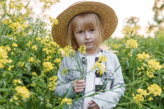 Blonde girl in a field with yellow flowers. A girl in a straw hat is picking flowers in a field. A field with rapeseed