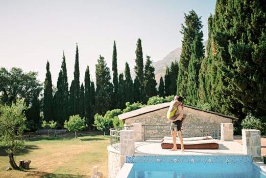 Man holds woman in his arms while standing by a pool deck chair. High quality photo