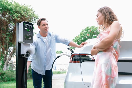 Happy and lovely couple with eco-friendly conscious recharging electric vehicle from EV charging station. EV car technology utilized as alternative transportation for future sustainability. Synchronos