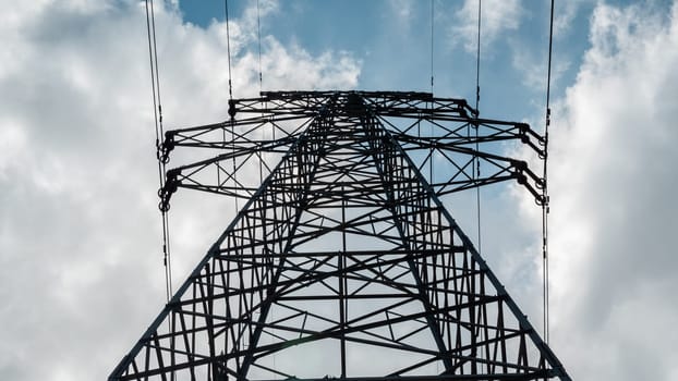 Bottom view of a high-voltage electricity pylon against blue sky with clouds at sunny day. High-voltage power transmission tower. Power engineering.