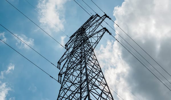 Bottom view of a high-voltage electricity pylon against blue sky with clouds at sunny day. High-voltage power transmission tower. Power engineering.