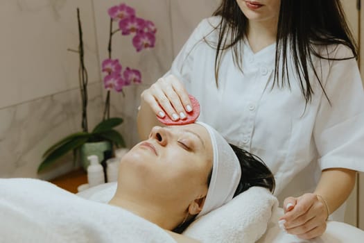 A young Caucasian girl cosmetologist cleanses the face of an adult female client using a pink scrubbing sponge, which lies on a massage table in a beauty salon, close-up side view.