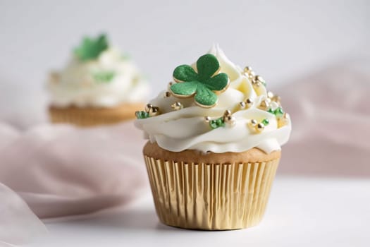 Small cupcake with cream and gold beads and a green clover on a light background. Green four-leaf clover symbol of St. Patrick's Day. A joyous time of celebration in the green color.