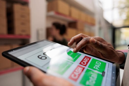 Worker checking online customers orders using tablet computer, preparing cardboard boxes before shipping. African american manager working at products inventory in warehouse. Close up concept